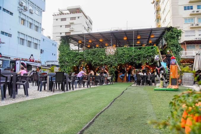 Restaurante al aire libre en una zona urbana, con mesas y sillas para los comensales, una terraza cubierta con luces cálidas y una fachada decorada con abundante vegetación. Se observan edificios alrededor y esculturas decorativas junto al área de césped artificial.